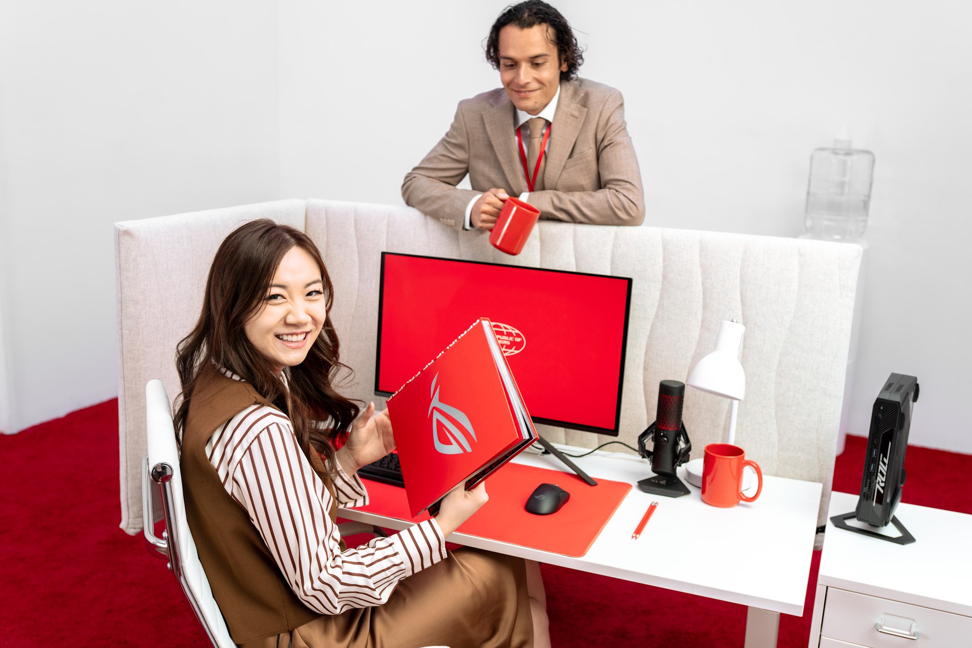 Một người phụ nữ ngồi ở bàn văn phòng cầm một cuốn sách ROG trong khi một chàng trai trẻ với cốc cà phê dựa vào bức tường phân chia của tủ